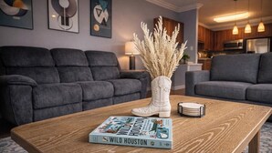 Uma sala de estar moderna com sofá, mesa de centro com um livro e objetos decorativos, e uma cozinha ao fundo.