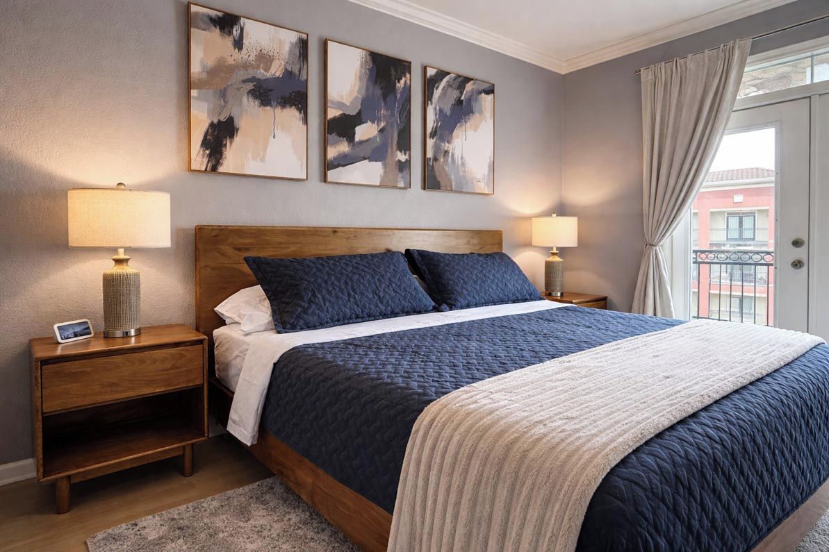 A bedroom with a wooden headboard, a bed with dark and light bedding, two bedside lamps, and abstract wall art.