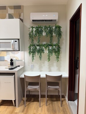 A compact kitchen with a microwave, a coffee machine, and a small refrigerator. There is a white desk with two chairs and a wall-mounted air conditioner.