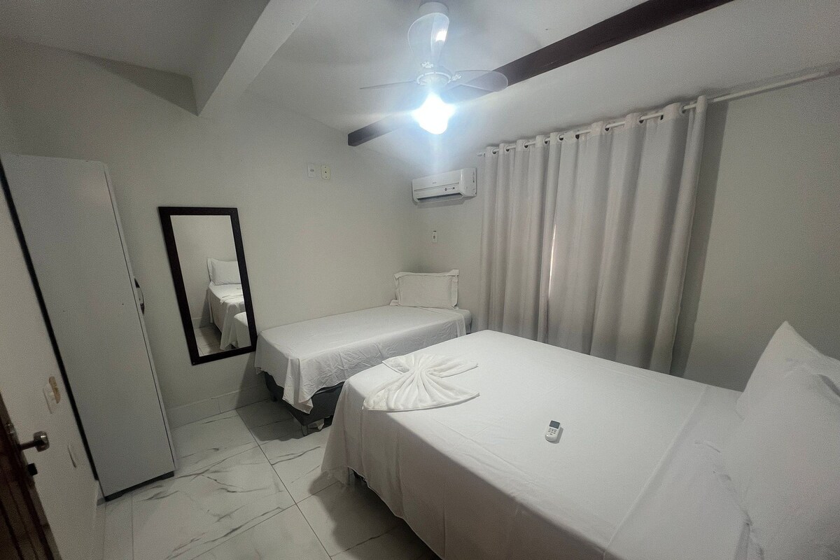 Quarto de hotel com duas camas de solteiro, ventilador de teto, ar condicionado, espelho e porta.