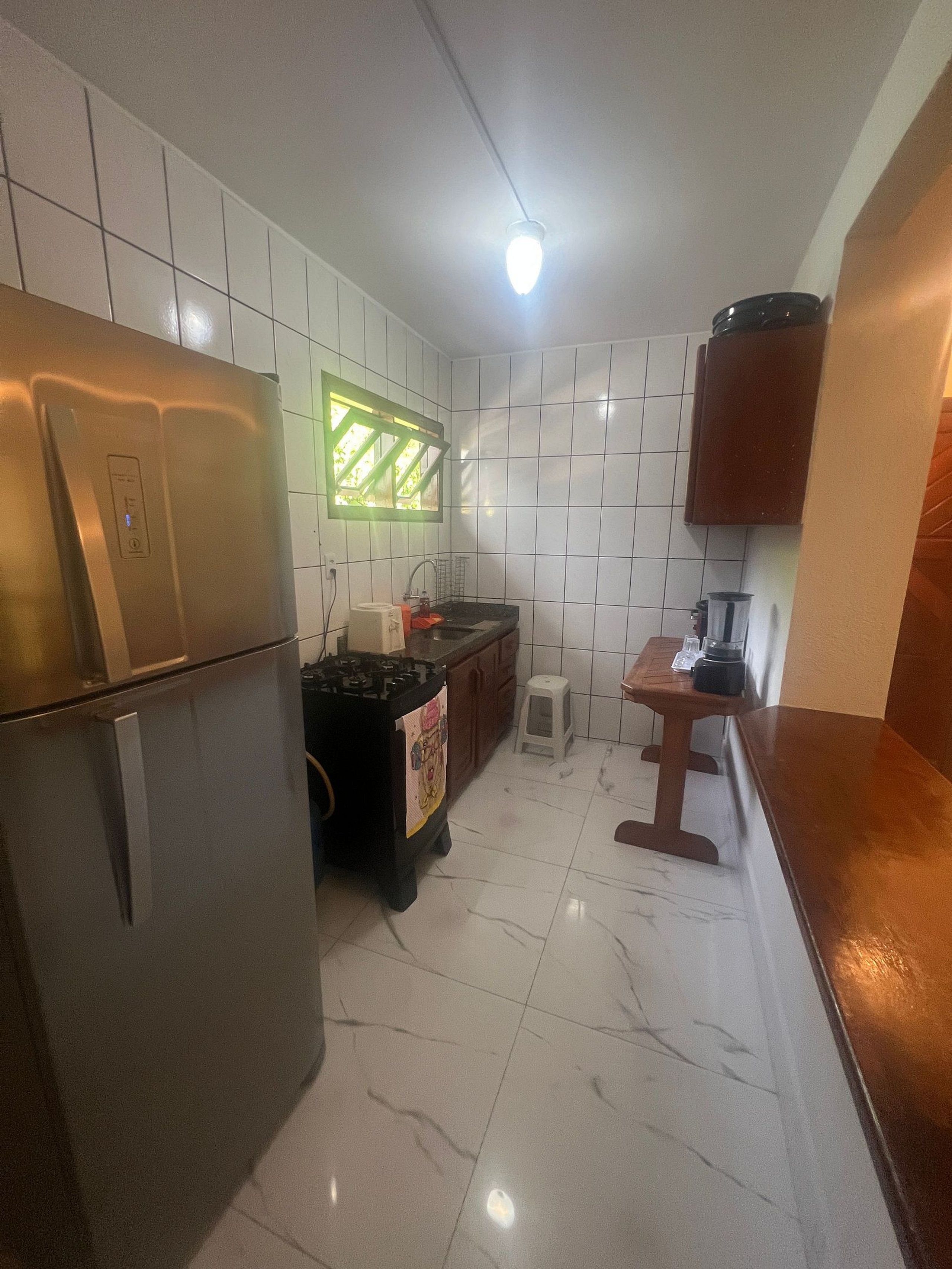Cozinha compacta com azulejos brancos, geladeira de aço inoxidável, pia, um banquinho pequeno e uma mesa de madeira.