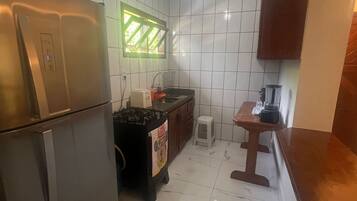 Cozinha compacta com azulejos brancos, geladeira de aço inoxidável, pia, um banquinho pequeno e uma mesa de madeira.