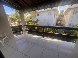 Un balcón con piso de baldosas, baranda de madera y vista a un edificio de fachada blanca y escalera.