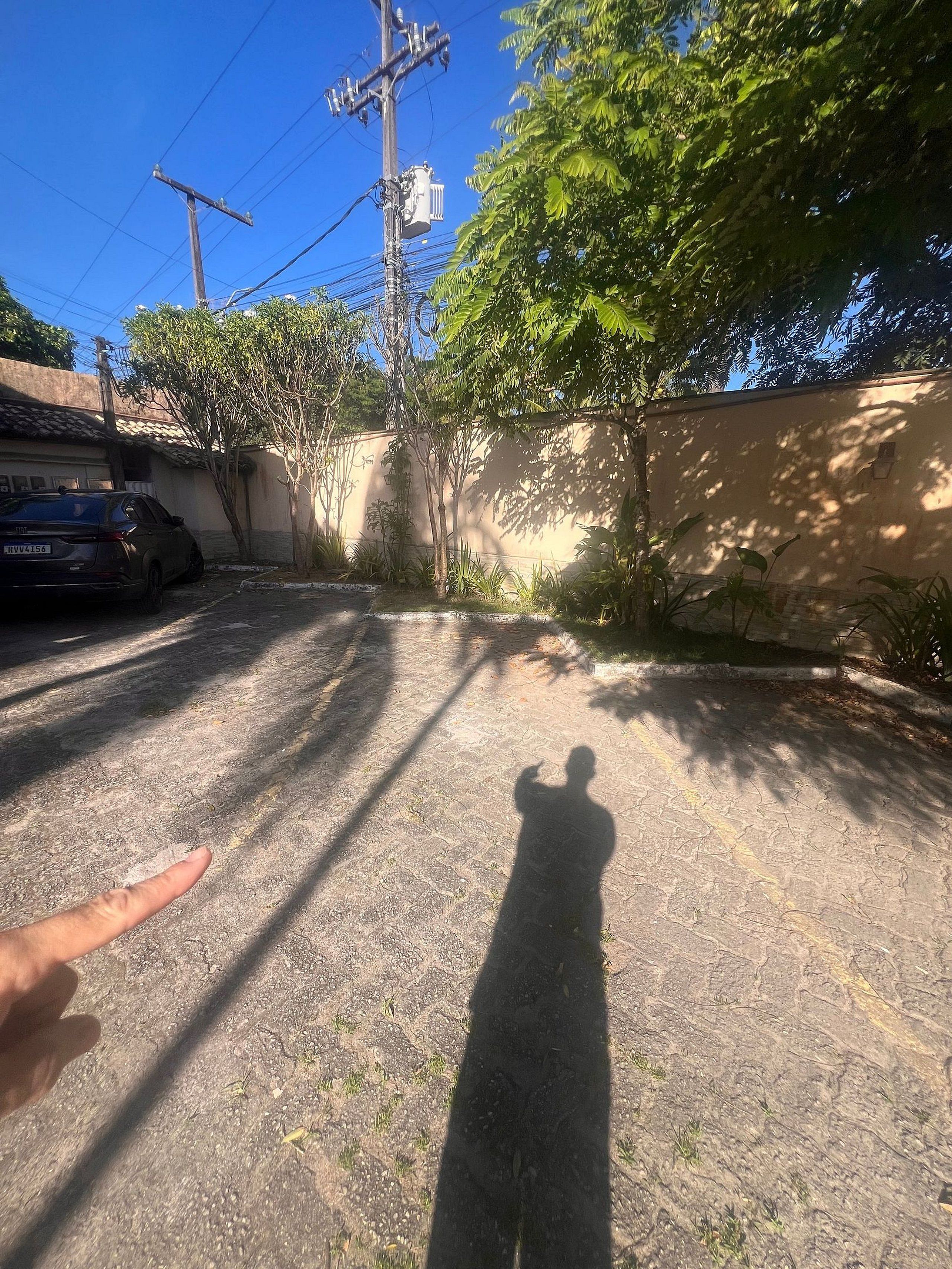Schatten einer Person auf einer gepflasterten Fläche, ein Auto parkt links, ein Strommast und eine Wand mit Vegetation im Hintergrund.