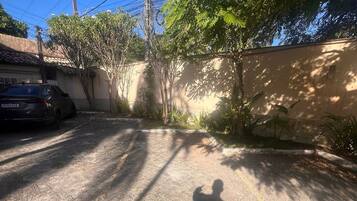Schatten einer Person auf einer gepflasterten Fläche, ein Auto parkt links, ein Strommast und eine Wand mit Vegetation im Hintergrund.