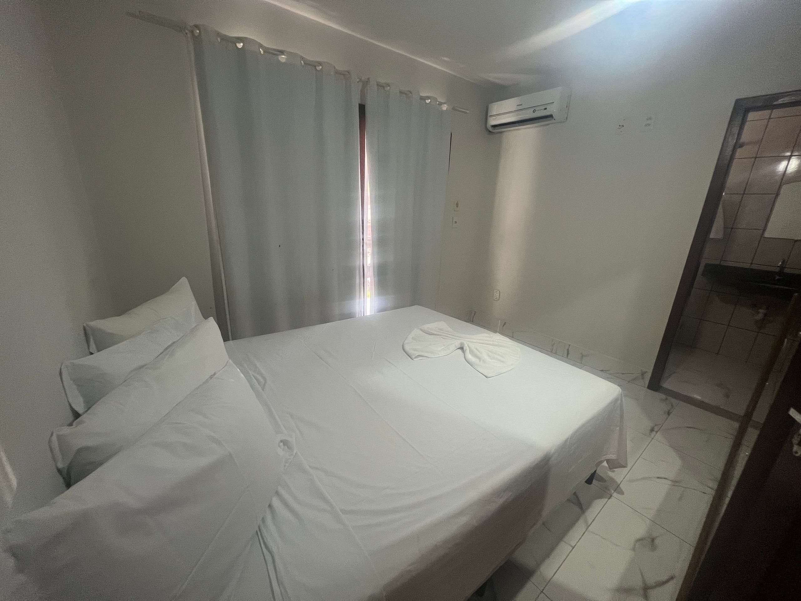 Une chambre d’hôtel avec un lit, des rideaux, un appareil de climatisation et une salle de bain visible à travers une porte en verre.