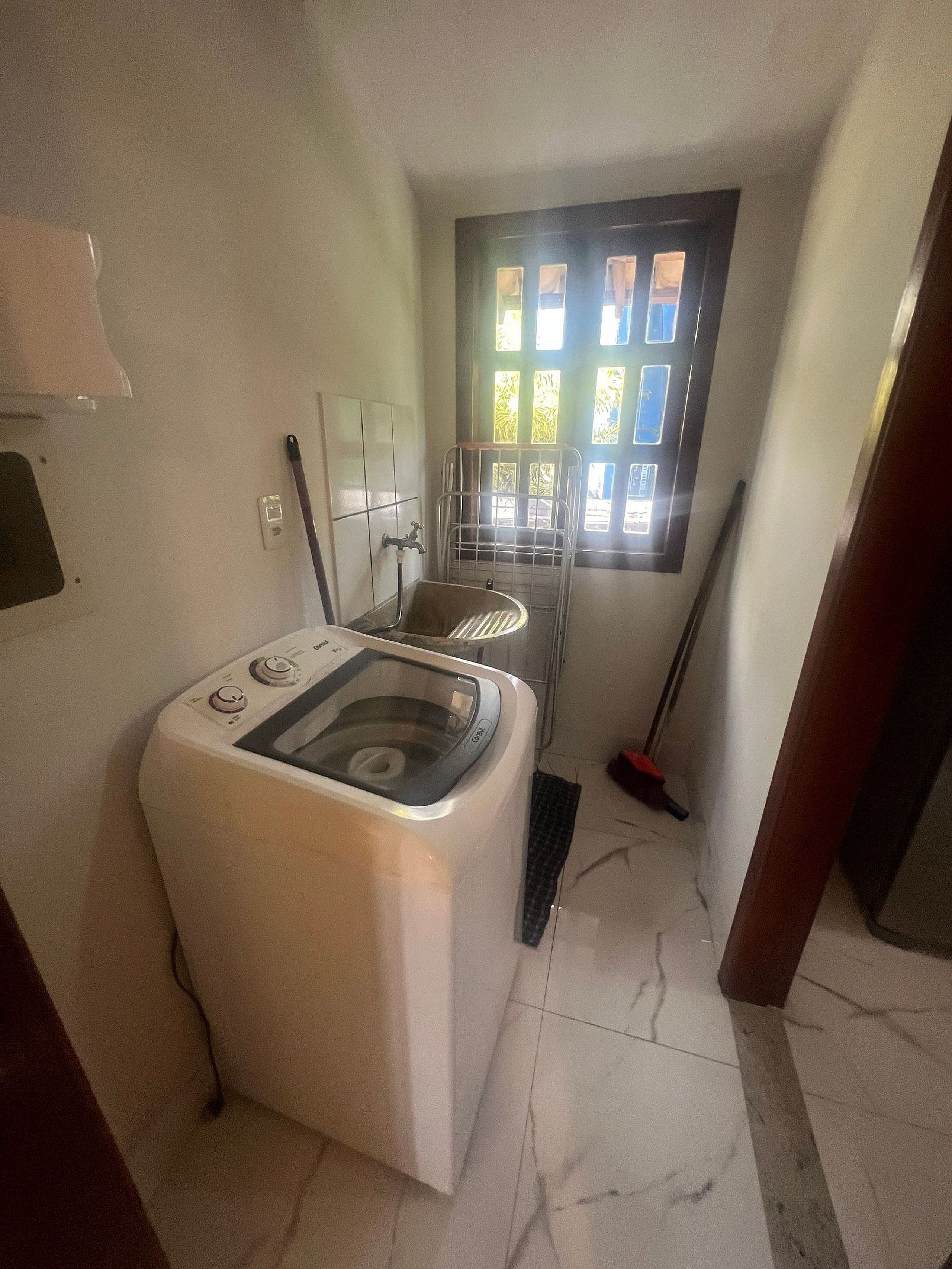 A laundry area with a washing machine, a window with blinds, and a broom.