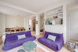 Un salon avec deux canapés violets, une petite table verte et une bibliothèque contenant des livres et des objets décoratifs.