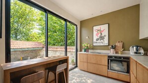 A modern kitchen with a large window, wooden cabinets, a built-in oven, and a dining area with two chairs. - La Maison Raspail - Welkeys (Bois-Colombes)