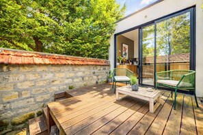 Een houten terras met een stenen muur, twee rieten stoelen en een tafel met een bloempot.