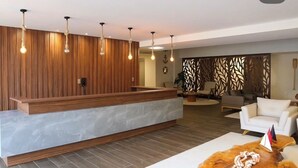 A reception area with a wooden reception desk, a white sofa, a decorative table with a flag, and a patterned wall. - ORA BEACH RESIDENCE  (Ipojuca)