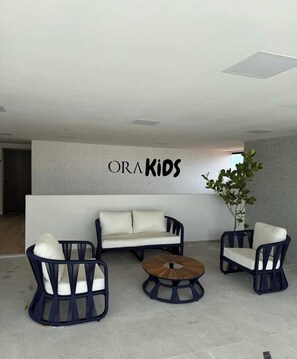 A modern lounge area with a sofa, two armchairs, and a coffee table. A wall-mounted sign reads 'ORA KIDS'. - ORA BEACH RESIDENCE  (Ipojuca)