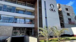 A multi-story building with a prominent logo on the facade, a covered entrance, and a landscaped area with palm trees. - ORA BEACH RESIDENCE  (Ipojuca)