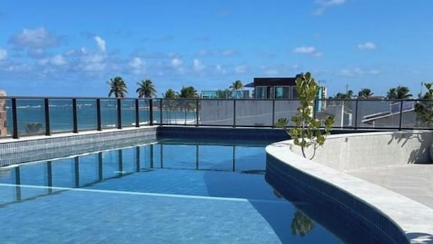 A swimming pool with a curved edge, surrounded by a concrete deck and a metal fence. - ORA BEACH RESIDENCE  (Ipojuca)