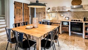 Ein Holzespeisesaal mit Stühlen, eine Küche mit Herd, Backofen und hängendem Kochgeschirr sowie Holzläden an den Fenstern.