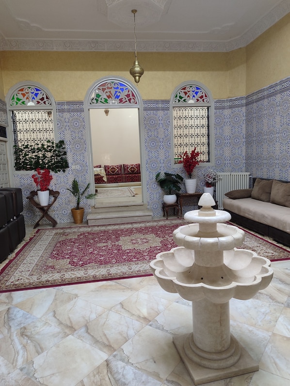 A room with a marble floor, a central fountain, a sofa, a coffee table, and a staircase leading to an upper level.