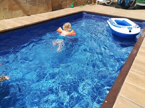 Une piscine avec un enfant dans l’eau, un flotteur bleu gonflable et un mur en pierre.