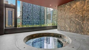 A modern indoor pool area with a glass wall, a marble wall, and a view of surrounding buildings. - LaneStay - 2BR Panoramic Views w Pool, Spa & Gym (Melbourne)