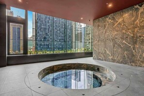 A modern indoor pool area with a glass wall, a marble wall, and a view of surrounding buildings. - LaneStay - 2BR Panoramic Views w Pool, Spa & Gym (Melbourne)