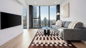 A modern living room with a large flat-screen TV, a grey sofa, a patterned rug, and a city view through large windows. - LaneStay - 2BR Panoramic Views w Pool, Spa & Gym (Melbourne)