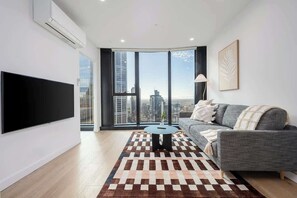 A modern living room with a large flat-screen TV, a grey sofa, a patterned rug, and a city view through large windows. - LaneStay - 2BR Panoramic Views w Pool, Spa & Gym (Melbourne)