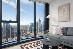 A modern living room with a large window offering a city view, a grey sofa, a coffee table, and a framed artwork on the wall. - LaneStay - 2BR Panoramic Views w Pool, Spa & Gym (Melbourne)