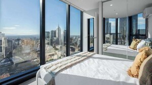 A modern hotel room with a large bed, a view of the cityscape, and a glass wall. - LaneStay - 2BR Panoramic Views w Pool, Spa & Gym (Melbourne)