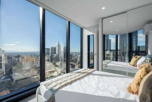 A modern hotel room with a large bed, a view of the cityscape, and a glass wall. - LaneStay - 2BR Panoramic Views w Pool, Spa & Gym (Melbourne)