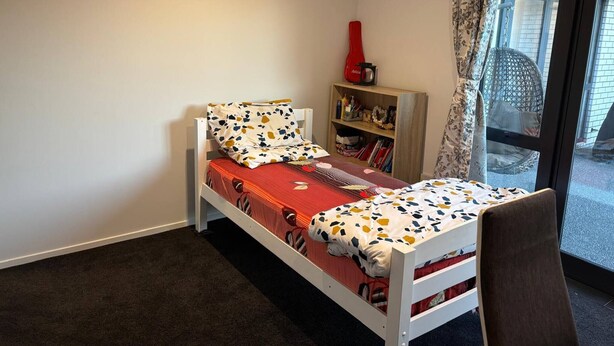 Una cama individual con una colcha estampada, una pequeña estantería con libros y un objeto rojo, y vista al exterior a través de un ventanal.