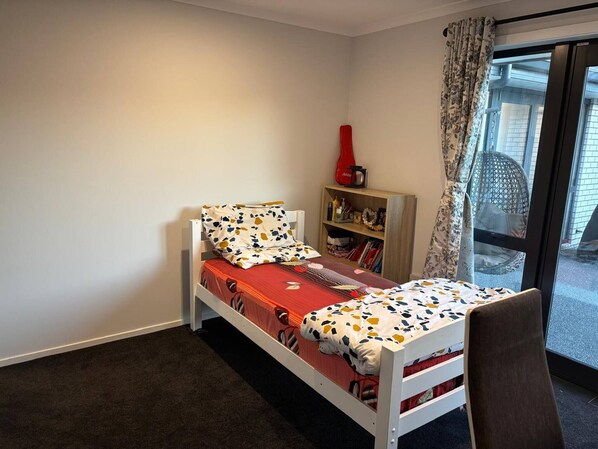 A single bed with a patterned bedspread, a small bookshelf with books and a red object, and a view of the outside through a glass door.