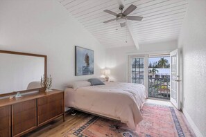Une chambre à coucher comprenant un lit, une commode, un ventilateur de plafond, un tableau, une lampe et une vue sur l’extérieur, accessible par une porte coulissante.