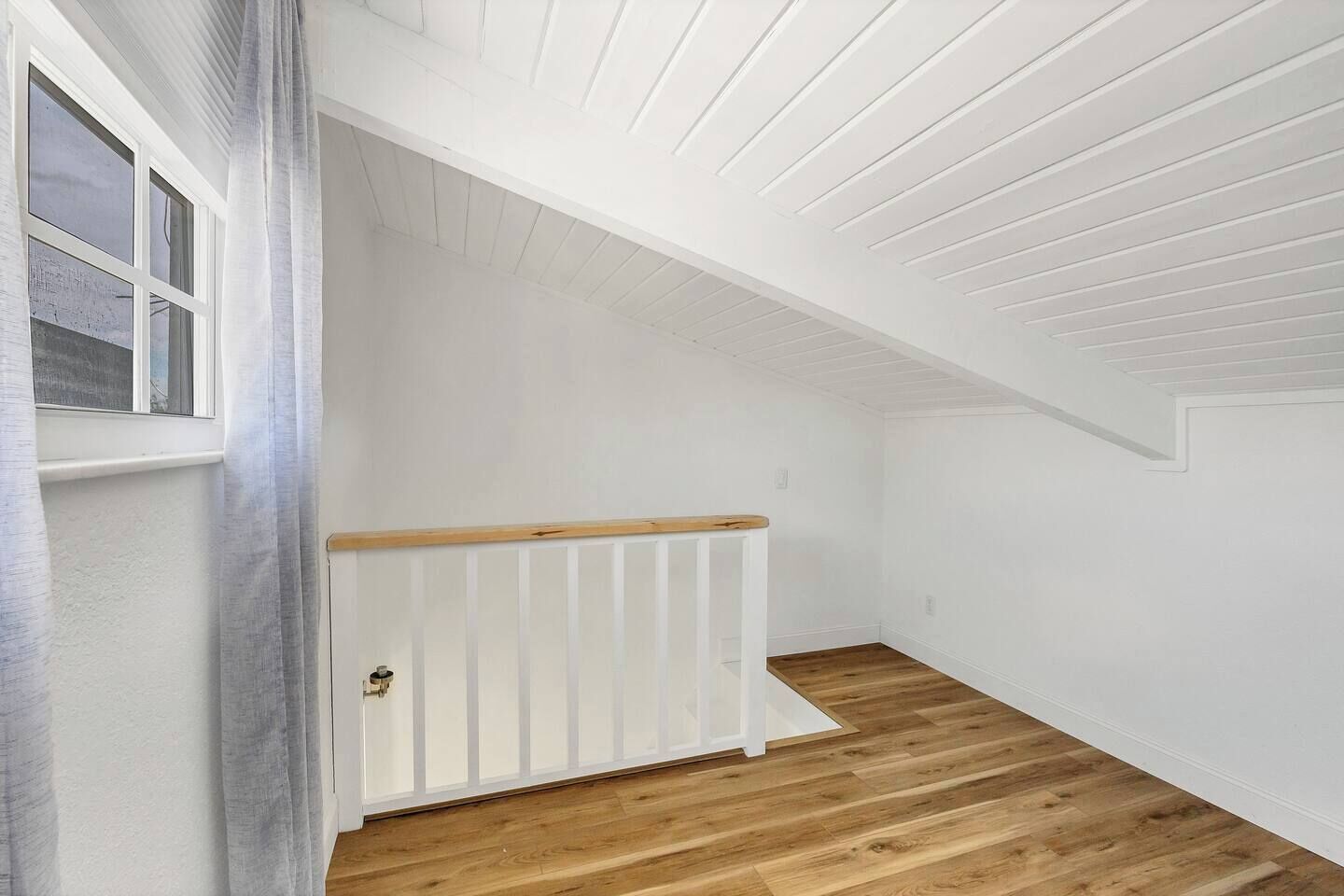 A room with a sloped ceiling, wooden flooring, a white radiator, and a window with curtains.
