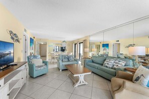 A spacious living room with a large TV, multiple sofas, a dining area, and a kitchen in the background. - Gorgeous Cocoa Beach Condo with Private Balcony Heated Pool & Hot Tub! ☀️ (Cocoa Beach)