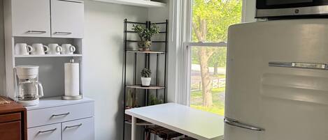 Una cocina con un refrigerador blanco, una cafetera, una mesa blanca y una ventana con vistas a árboles.