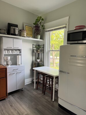 Eine Küche mit einem weißen Kühlschrank, einer Kaffeemaschine, einem weißen Tisch und einem Fenster mit Blick auf Bäume.