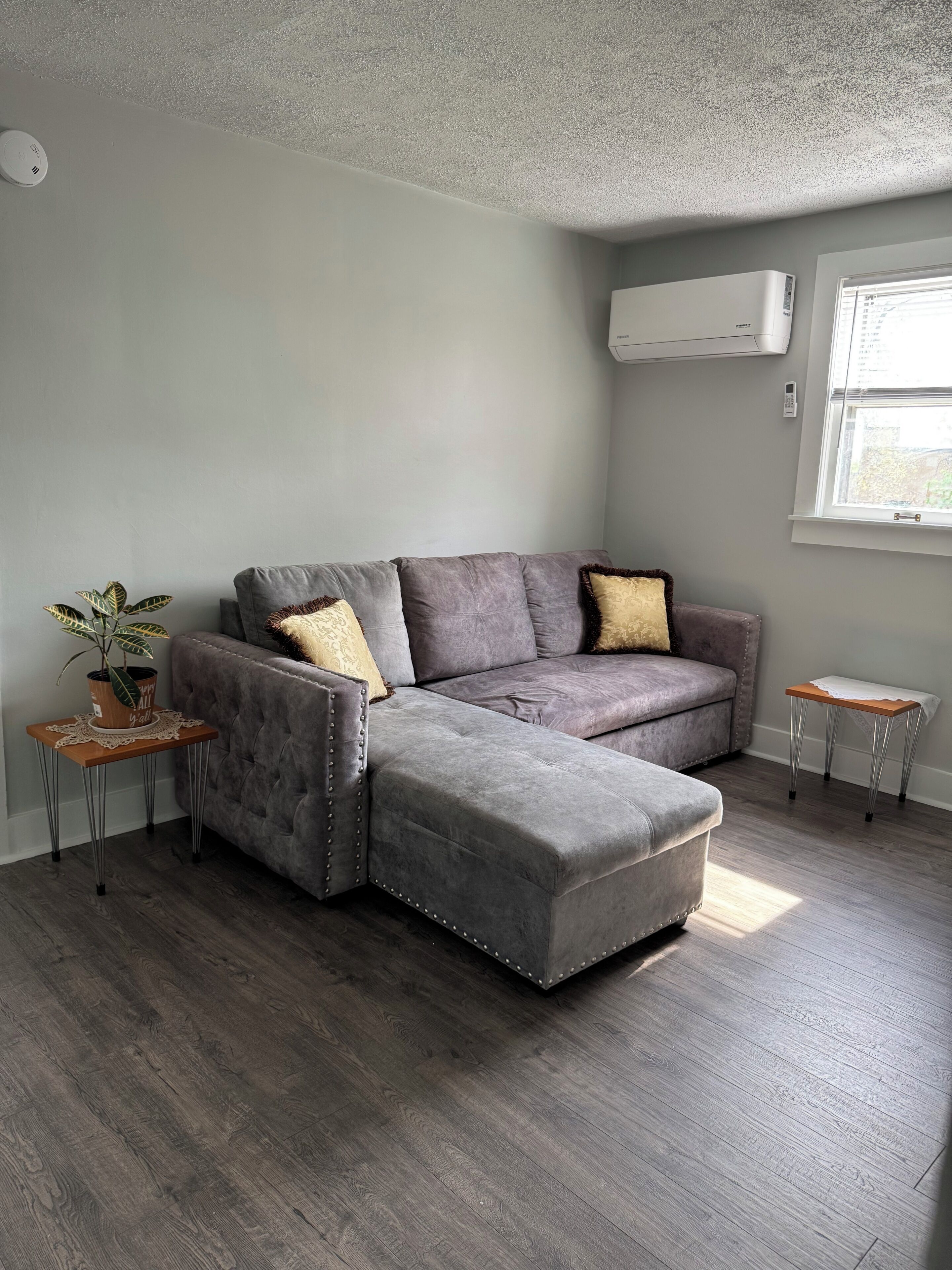 A living room with a sectional sofa, a small side table with a plant, a window with blinds, and an air conditioning unit on the wall.