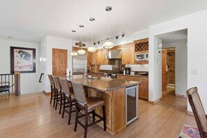 A modern kitchen with wooden cabinets, a central island with bar stools, stainless steel appliances, and pendant lighting. - Luxury Timbers Townhome | Walk to Town & Ski (Ketchum)
