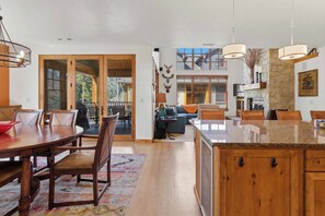 A spacious living area with a large wooden dining table, comfortable seating, a fireplace, and a view of the outdoors. - Luxury Timbers Townhome | Walk to Town & Ski (Ketchum)