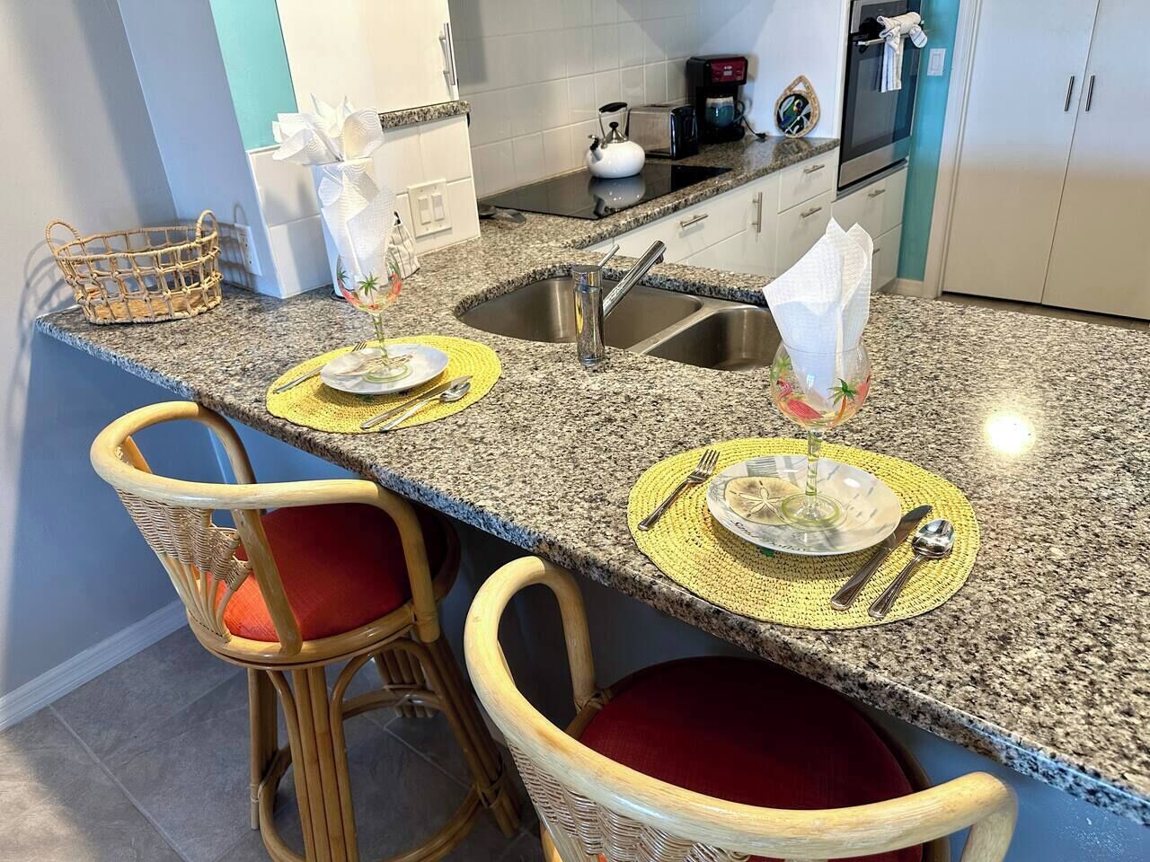 A kitchen with granite countertops, two wicker chairs with red cushions, a sink, and a coffee maker.