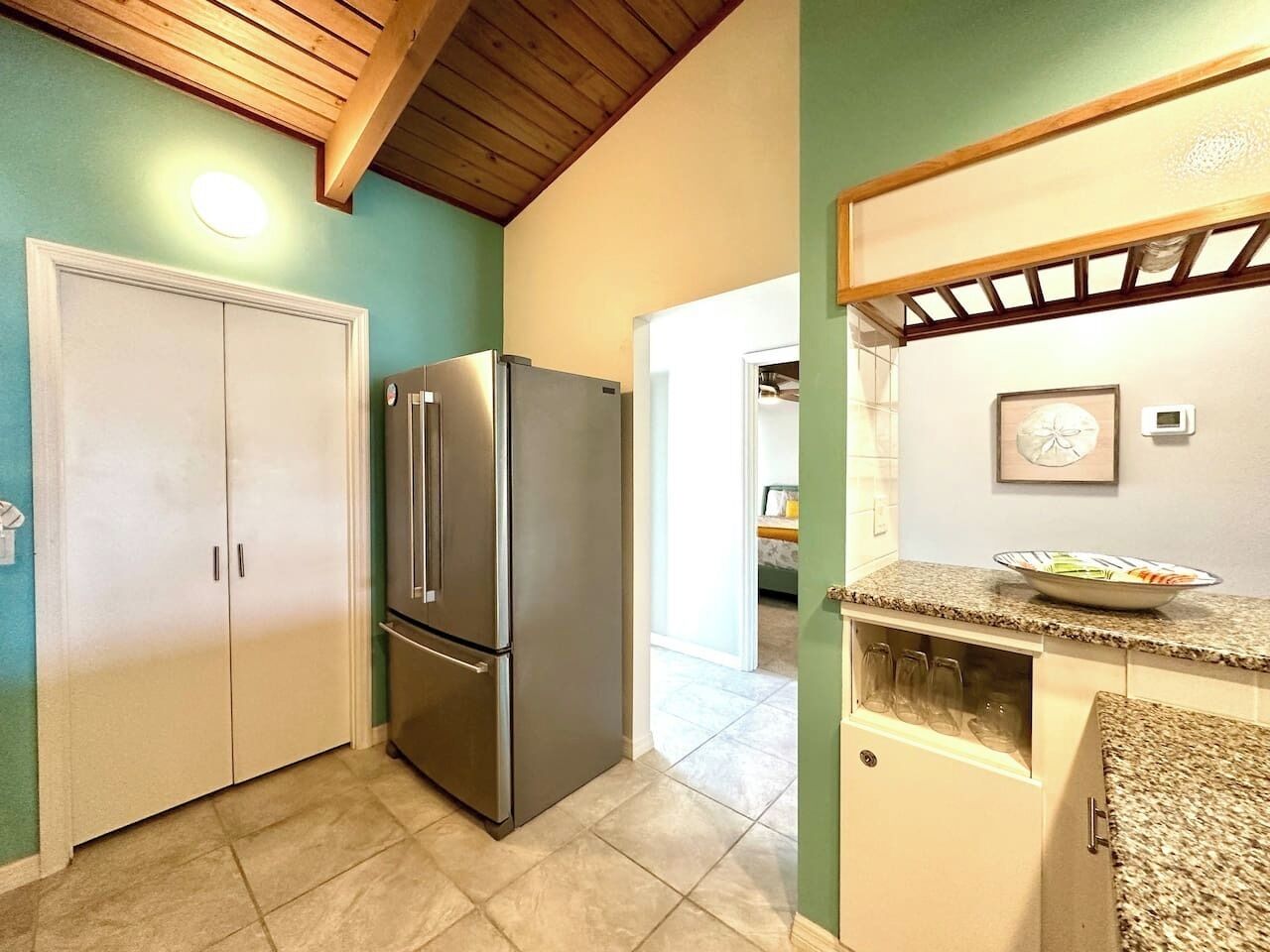A kitchen with a double-door refrigerator, a countertop with a bowl of fruit, and a wooden ceiling.
