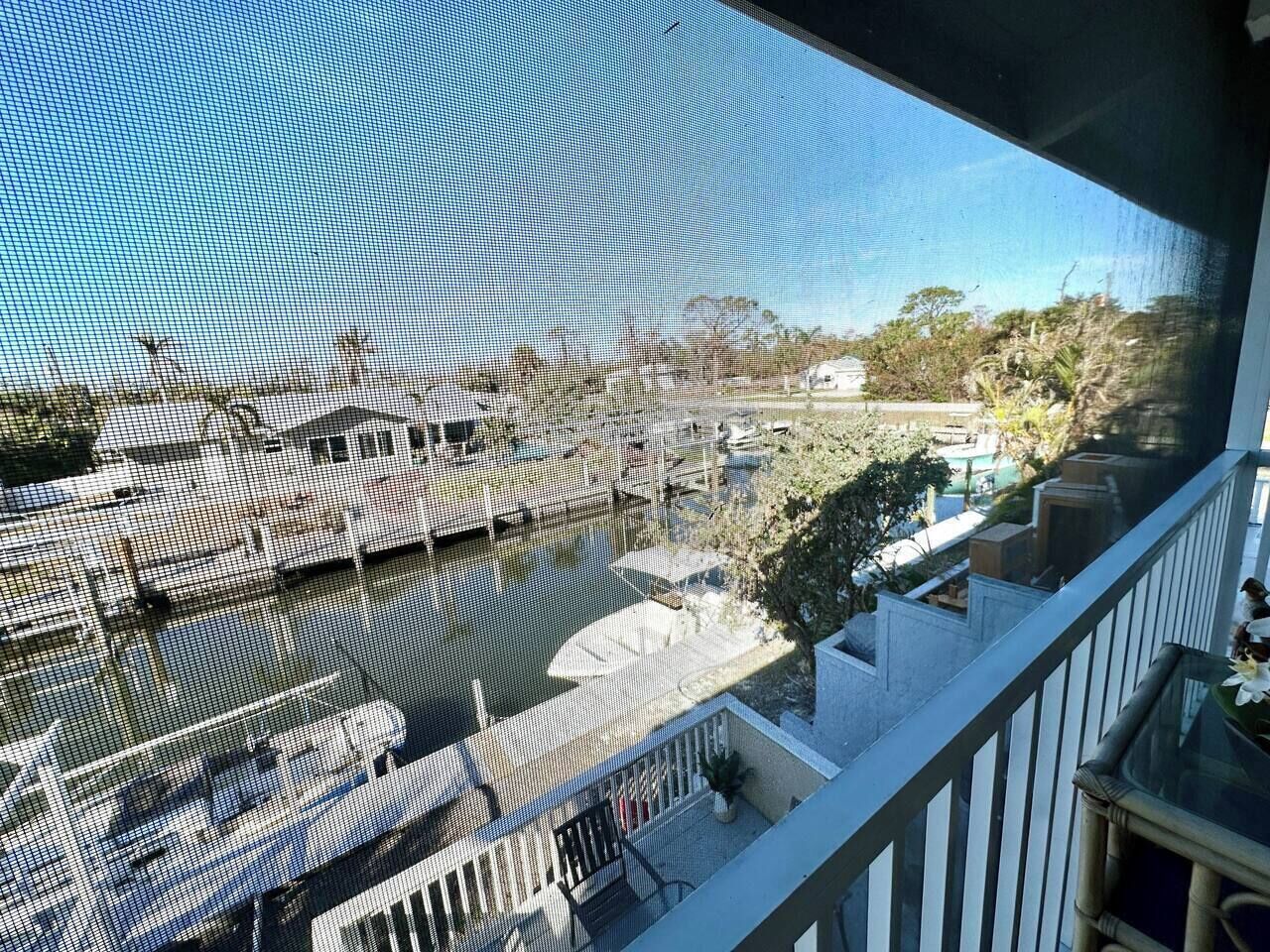 A view through a window of a marina with boats docked, a balcony with a railing, and some greenery in the background.