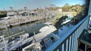 Une vue à travers une fenêtre donnant sur une marina où sont amarrés des bateaux, un balcon avec une balustrade, et, au loin, quelques éléments de végétation.