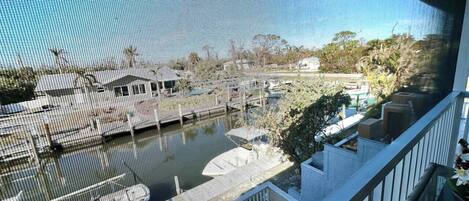 A view through a window of a marina with boats docked, a balcony with a railing, and some greenery in the background.