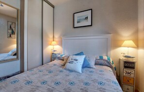 A bedroom with a bed, two bedside lamps, a framed picture, and a nightstand with a sign. - Appartement proche plage (Saint-Georges-de-Didon)