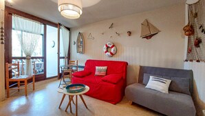 A living room with a red sofa, a grey sofa, a small round table with a plate, and a chair. There are wall decorations including a life ring, a ship, and a bird.