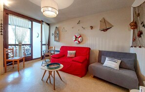 A living room with a red sofa, a grey sofa, a small round table with a plate, and a chair. There are wall decorations including a life ring, a ship, and a bird. - Appartement proche plage (Saint-Georges-de-Didon)