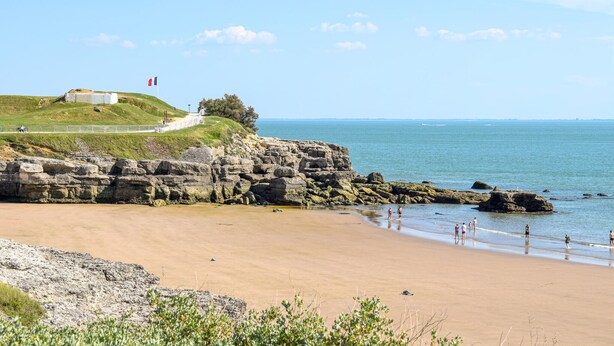 A coastal landscape with a sandy beach, rocky cliffs, and a grassy hill with a flag.