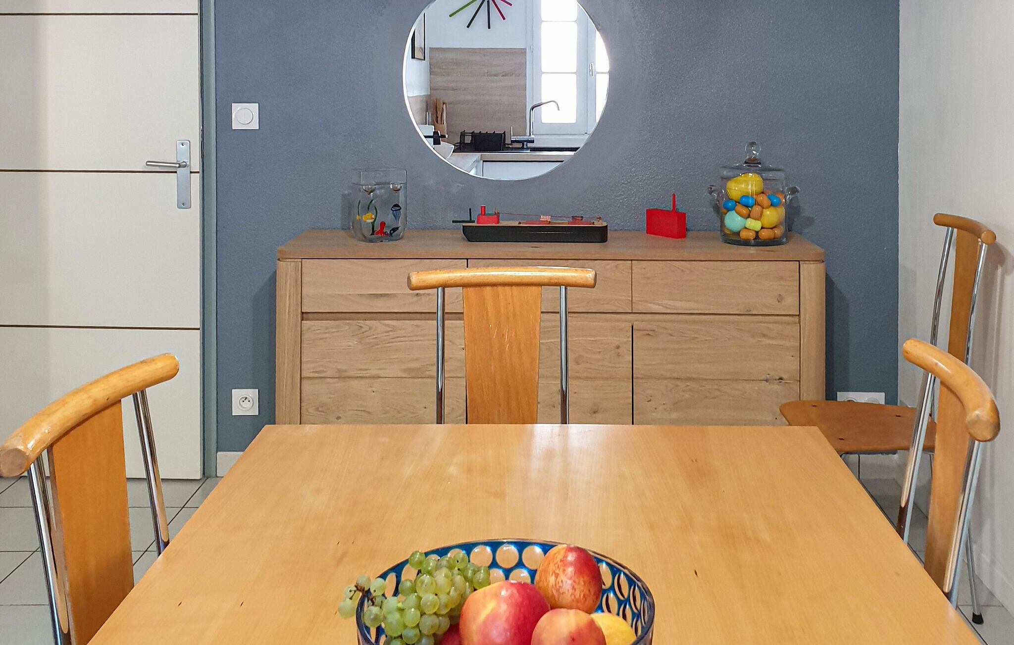 A dining area with a wooden table, chairs, a bowl of fruit, a mirror, and a kitchen counter with various items.