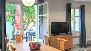 A dining area with a table, chairs, a fruit bowl, a television, and a view of greenery outside.