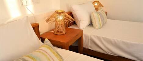 Una habitación con dos camas, cada una con cubrecama blanco y almohadas a rayas. Hay dos mesitas de noche de madera con lámparas, y se observa un enchufe en la pared.